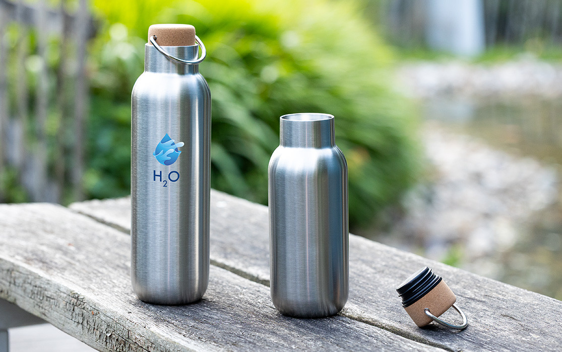 Gourdes en acier inoxydable REFLECTS® avec bouchon en liège – un design durable pour vos déplacements. Deux gourdes en acier inoxydable argenté avec bouchon en liège posées sur un banc en bois à l'extérieur, l'une d'elles arborant le logo H2O.