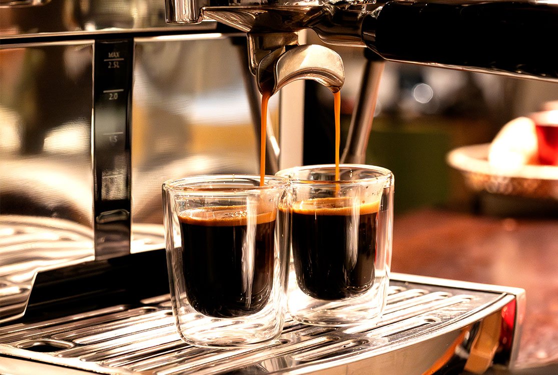 Zwei Espressobecher aus Glas unter der Kaffeemaschine mit Espresso darin