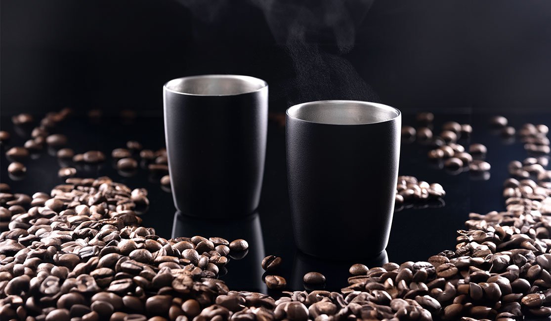 Zwei Espressobecher mit heißem Espresso darin und Kaffeebohnen rund herum
