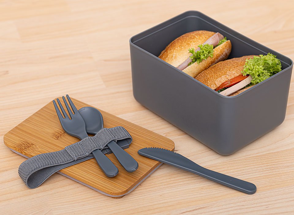 Lunchbox mit zwei belegten Brötchen darin und dem Besteck-Set auf dem Bambusdeckel 